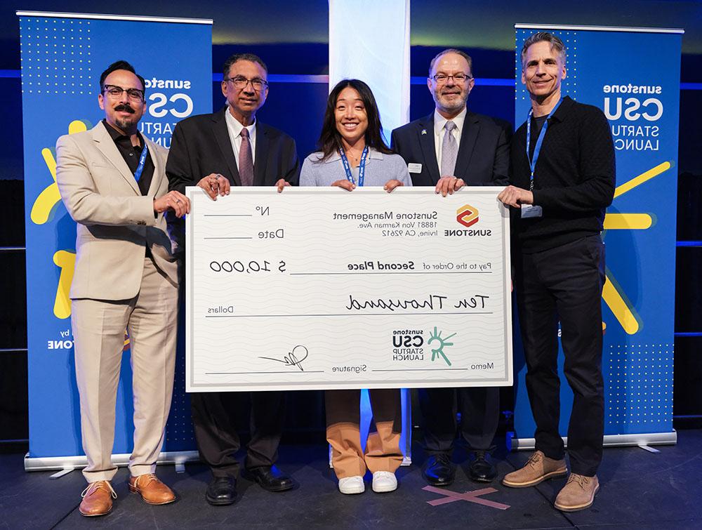 A female, college-age startup founder poses onstage with two sponsors, two hosts, and a giant check for $10,000