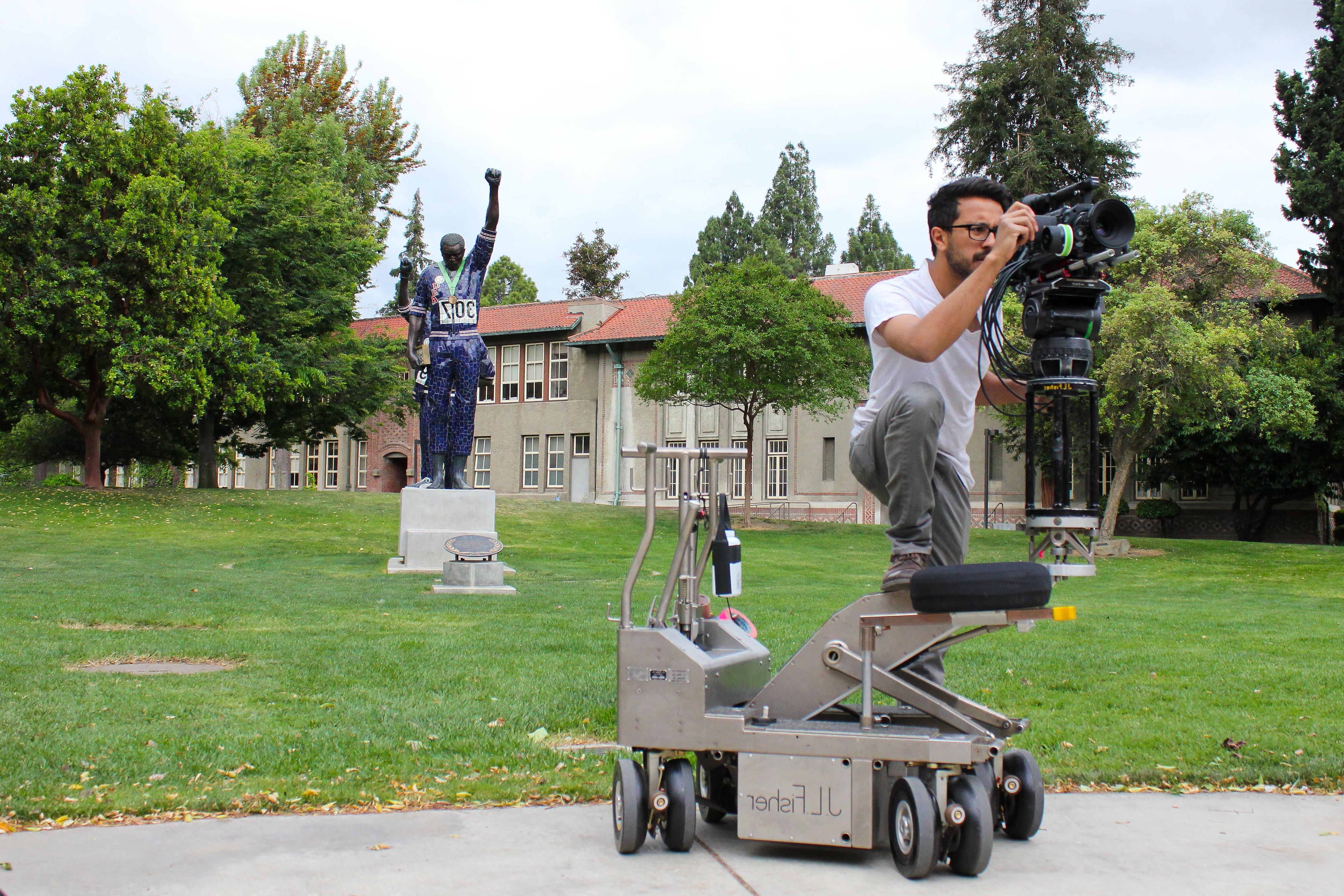 A student stands on a dolly with a camera while filming for a project.